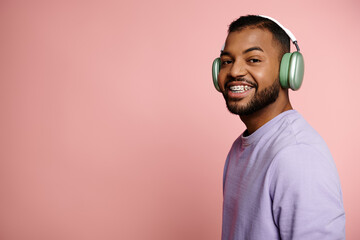 Young African American man with braces smiling brightly while wearing headphones on a pink...