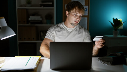 A focused young caucasian man with a beard works late at his office desk, holding a credit card.