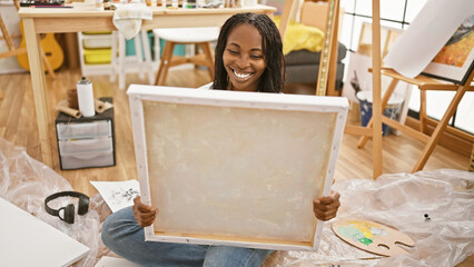 Smiling young woman artist holding a blank canvas in a creatively cluttered studio interior.