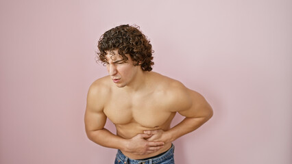 Young shirtless man with curly hair grimaces against a pink background, exuding pain or discomfort.