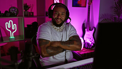 Handsome black man with beard and tattoos crosses arms in a neon-lit gaming room at night.