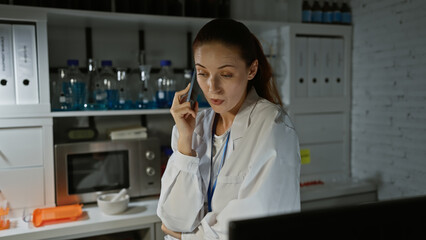 A thoughtful woman in a lab coat talks on the phone in an indoor laboratory setting.