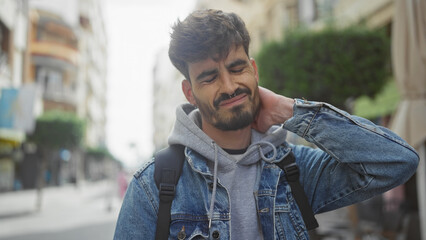 A young bearded hispanic man in casual attire on a city street, expressing neck pain.