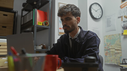 A thoughtful young hispanic man with a beard sits in a detective's office, surrounded by...