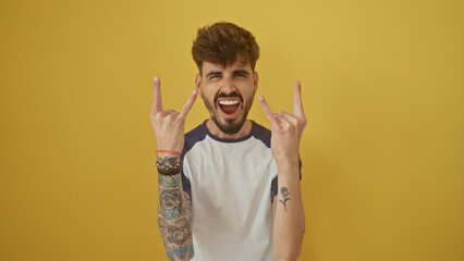 A bearded, tattooed man in casual clothing showing rock signs over a vibrant yellow background