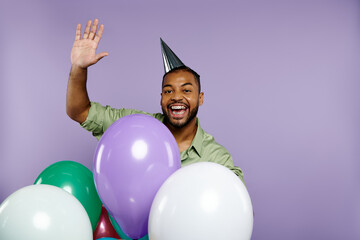 Young African American man in party hat smiling, holding colorful balloons against a purple background.