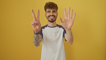 Happy young man with beard gesturing number seven with his fingers against a vibrant yellow...