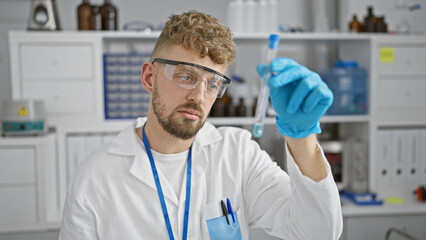 A young caucasian man with a beard, wearing safety glasses, examines a blue liquid in a test tube in a laboratory.