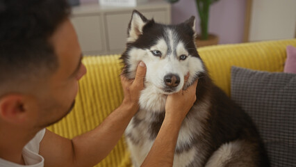 Young hispanic man affectionately pets his husky dog in a cozy living room setting, embodying companionship and domestic tranquility. © Krakenimages.com
