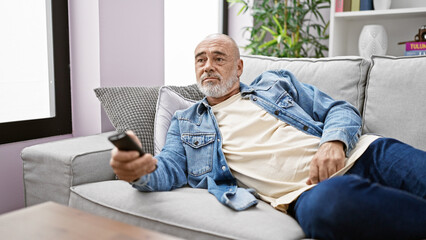 Mature man with beard in denim at home on couch holding remote