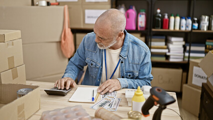 A senior hispanic man with a beard and bald works indoors, using a calculator near cardboard boxes, with euro currency visible.