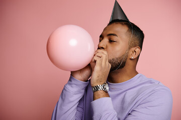 Young African American man wearing braces is happily blowing up a balloon while wearing a colorful...