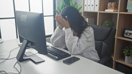 Stressed female doctor in hospital office covers face with hands, showcasing burnout and fatigue at...