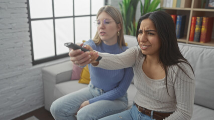 Two women sitting on a couch in a living room, engaged in watching television with a remote in hand.