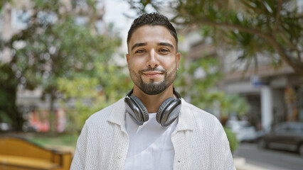 A handsome man with headphones smiling outdoors in a green city environment.