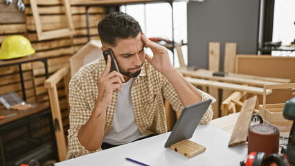 A stressed man talks on the phone in a cluttered carpentry workshop while looking at a tablet.