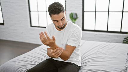 Handsome man in white examining wrist pain while sitting thoughtfully on bed in a bright, modern...