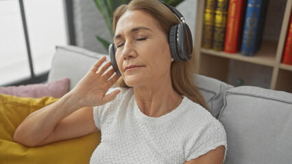 Mature woman enjoying music with headphones while relaxing on a sofa in a cozy living room.