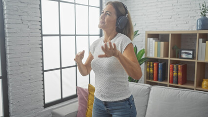 A middle-aged woman enjoys listening to music on headphones while dancing alone in a bright, modern...