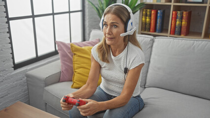 Mature woman playing video games with a controller and headset while seated on a couch indoors.