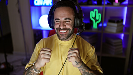 A beaming hispanic man wearing headphones enjoys gaming at home in a dark room lit by colorful led...