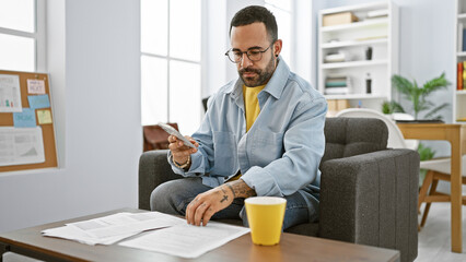 Handsome hispanic man with beard wearing glasses, comfortably working on finances in a sunny modern...