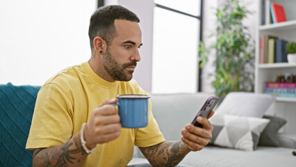 Handsome hispanic man with beard using smartphone and enjoying coffee in modern living room