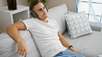 A relaxed young hispanic man wearing headphones lounges on a couch indoors, conveying ease and...