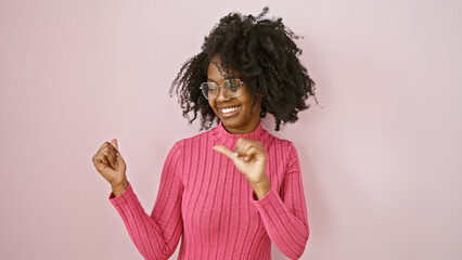 African american woman pointing sideways in a pink sweater and glasses against a plain background.