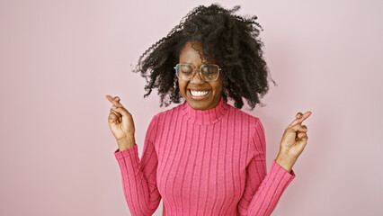 A cheerful black woman with glasses smiles while crossing her fingers in an indoor pink setting.