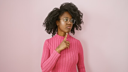 African american woman wearing glasses and pink sweater contemplates indoors against a plain...