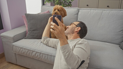 A middle-aged hispanic man and his poodle are taking a selfie in the living room of a modern...