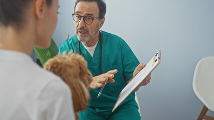 Hispanic veterinarian consults with a woman holding a poodle inside a clinic room.