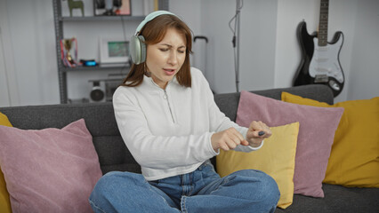 Obraz premium A displeased young woman with headphones sitting on a sofa holding a remote in a modern living room