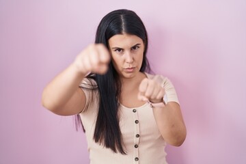 Fototapeta premium Young hispanic woman standing over pink background punching fist to fight, aggressive and angry attack, threat and violence