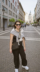 Fototapeta premium Smiling young woman wearing sunglasses stands on a budapest street with the st. stephen's basilica in the background.