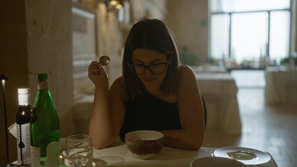 Young hispanic woman enjoying a meal indoors at an italian restaurant, examining her food with a...