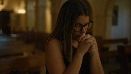 A beautiful young hispanic woman is seen praying inside a dimly lit christian church in italy, with...