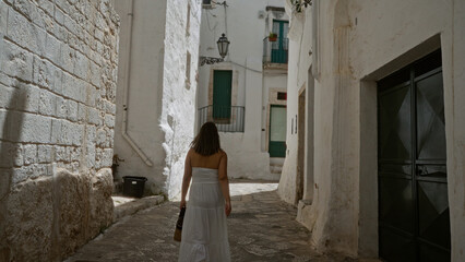 A young hispanic woman in a white dress explores the charming old town of ostuni, puglia, italy,...