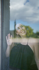 Hispanic young woman looking out of a window indoors, smiling beautifully with sunlight reflecting...