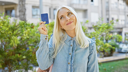 Confident young blonde woman joyfully holds credit card, deep in thought about her financial freedom while soaking up the sun in the park
