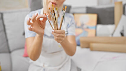Beautiful blonde young woman sitting relaxed on sofa, choosing paintbrush in indoor art studio