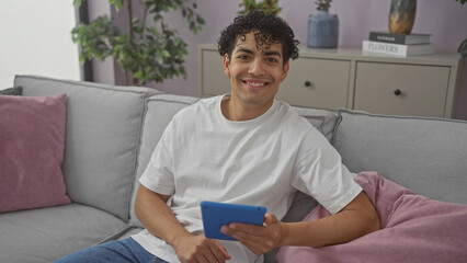 Smiling young hispanic man with tablet relaxing on living room sofa at home.