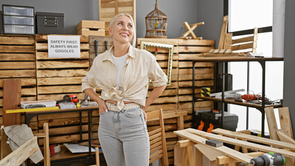 Vibrant young blonde female carpenter, smiling brightly and confidently, standing in her carpentry workshop, proud of her profession in the woodworking business