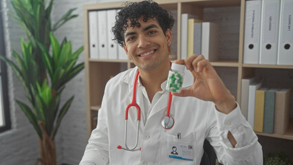 Smiling young hispanic male doctor holding medication bottle in a hospital office