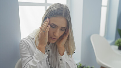 A young, blonde, caucasian woman in a white shirt holds her head in a clinic interior, reflecting...