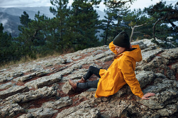 Solo Traveler in Yellow Raincoat and Hat Sitting atop Mountain Rock, Capturing the Beauty of Nature