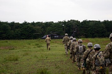 A group of soldiers are walking in a field with a helicopter in the background