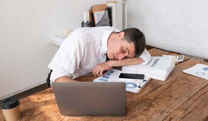 Obraz premium Businessman exhausted and asleep at desk with laptop and financial reports, concept of workplace fatigue and stress