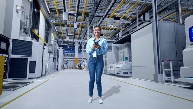 Full Body Of An Asian Female Professional Worker Standing With Her Smarphone at Factory Digitalization for Innovative Project, She Is Looking At The Camera With A Smile